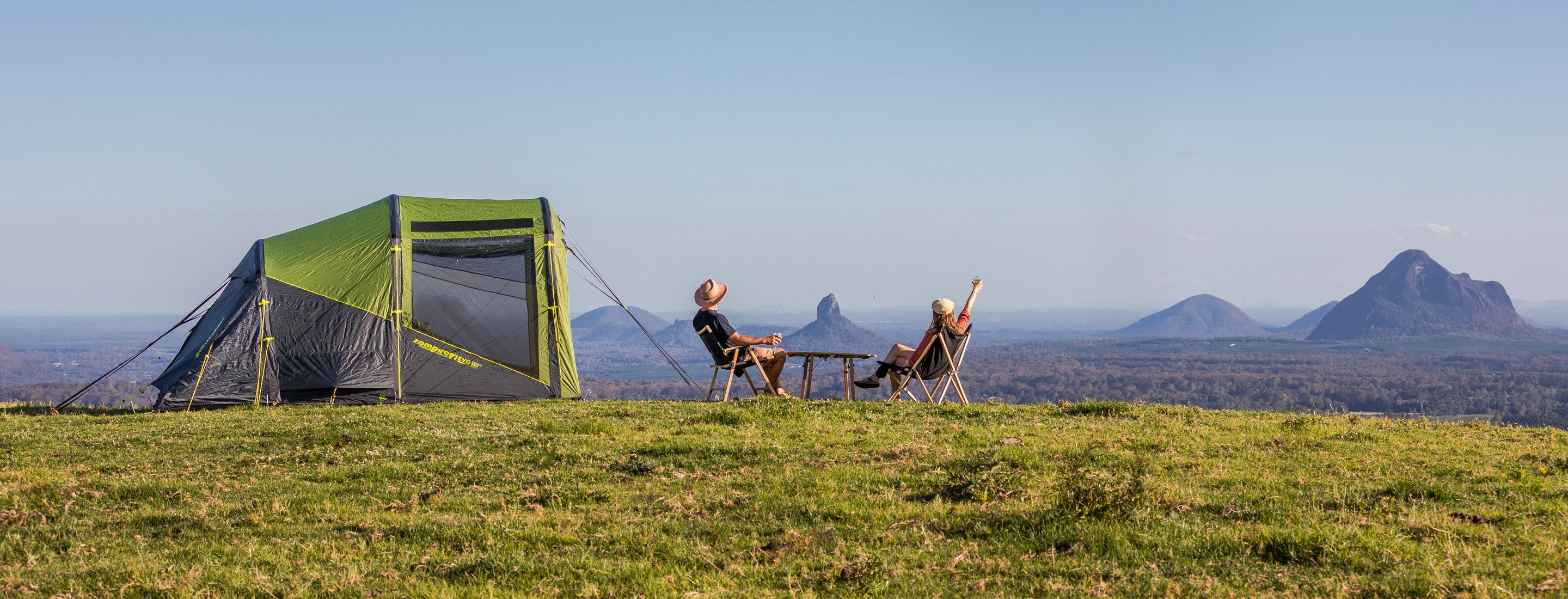 Two people sitting on a grassy hill with a green tent and scenic background