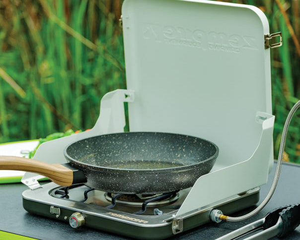 A portable camping stove with an open white lid, holding a black speckled frying pan. The stove sits on a dark tabletop outdoors with tall green reeds in the background.